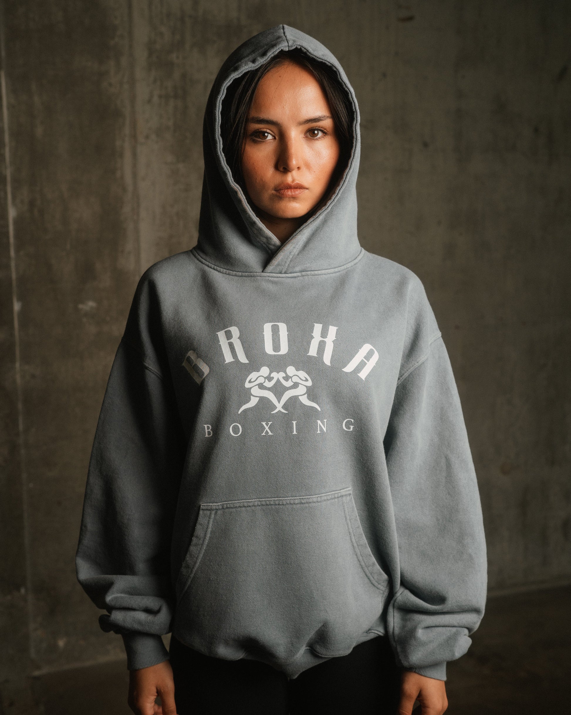 Person wearing a gray hoodie with Broxa Boxing' logo against a concrete wall.