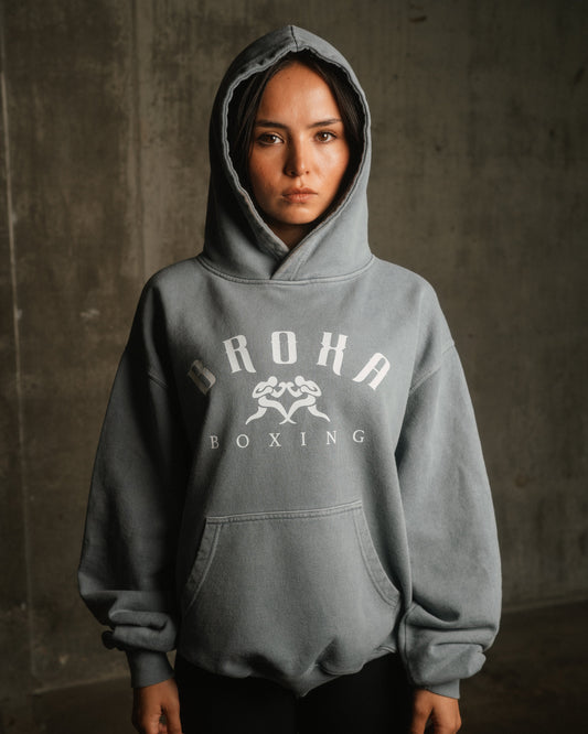 Person wearing a gray hoodie with Broxa Boxing' logo against a concrete wall.